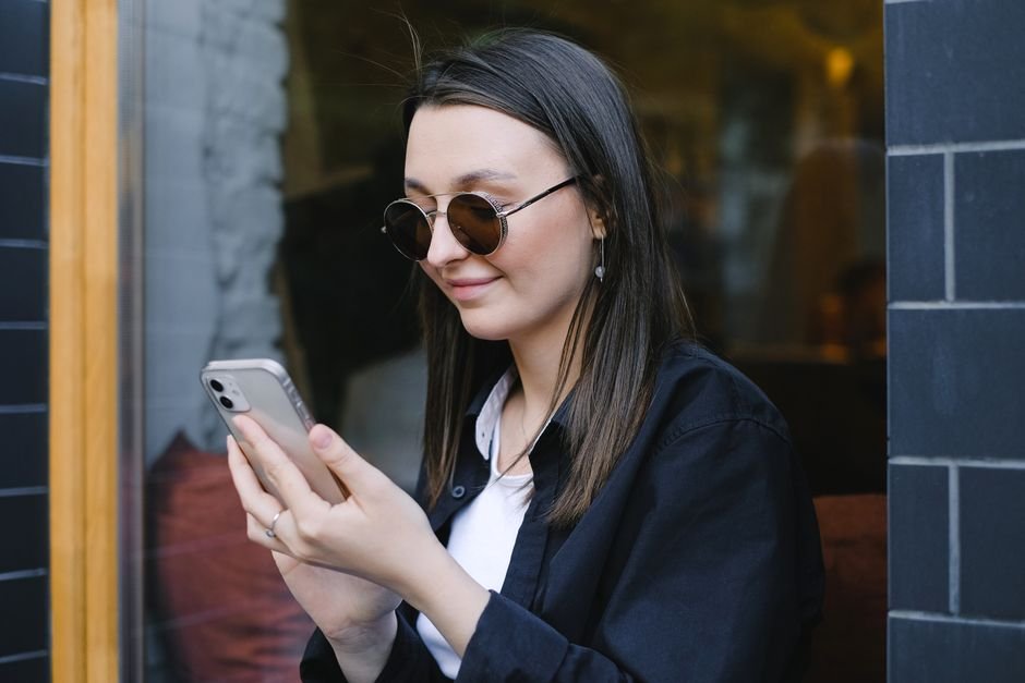 Une jeune femme en lunettes de soleil souriant devant son smartphone, en extérieur